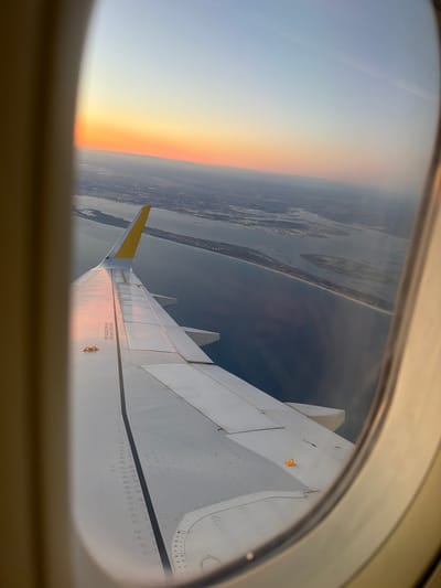 The image is an aerial view from an airplane window, looking out over a body of water and land. The setting sun casts a warm glow across the sky, transitioning from orange and yellow near the horizon to a pale blue higher up. An airplane wing, with a yellow tip, dominates the foreground. The wing has visible rivets and warning labels. Below, a coastline is visible, with a sandy beach meeting calm, dark blue water. Further inland, there are areas of land dotted with lights, suggesting a town or city. This view is consistent with an approach or departure from an airport near Alcácer do Sal, Portugal, likely over the Sado River estuary, which features a similar coastline and water system.