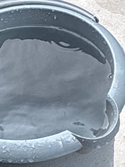 The image shows a close-up view of a gray bucket filled with water. The bucket appears to be made of plastic and has a textured surface. Water droplets are visible on the rim and sides of the bucket. The water inside is murky and reflects light in wavy patterns. The background is blurred and appears to be a light-colored, rough surface, possibly concrete or gravel. There are no people or animals visible in the image, and no specific actions or interactions are depicted. The scene is static and appears to be an outdoor setting. The specific location in Bad Hersfeld, Germany, is not identifiable from the visual information provided.
