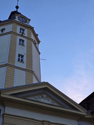 The image shows a close-up, low-angle view of a clock tower in Ansbach, Germany, against a clear blue sky. The tower, painted in cream and yellow, features several windows and architectural details. At the top of the tower, a clock face with Roman numerals is visible. A flagpole extends from the side of the tower. Below the tower, a section of a building with a triangular pediment and a carved coat of arms is visible. The overall scene is that of an historic building in an urban setting.
