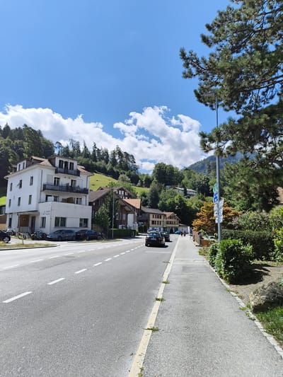 In Matten bei Interlaken, Switzerland, a road stretches through a village with buildings and a lush green hillside under a bright blue sky with scattered white clouds. On the left side of the road, a white, multi-story building with balconies and windows is visible, with bicycles and cars parked in front. Further down the street, a row of houses with red roofs and traditional architecture lines the road. Several cars are on the road, and a few people are walking on the sidewalk on the right. A tall lamppost with multiple signs stands near the sidewalk, and large trees, including a prominent pine tree on the right, provide shade. The environment is a mix of residential and natural elements, with trees, a hillside, and a paved road and sidewalk.