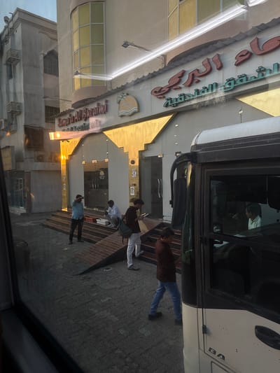 The image captures an exterior street scene in Al Buraimi Province, Oman, in front of the "Samara Al Buraimi Hotel Apartments." The building features a light-colored facade with golden decorative elements surrounding a recessed entrance, which includes glass doors and a set of tiered steps. An access ramp with a reddish-brown brick pattern is visible extending from the steps to the paved ground. The hotel's name, "Samara Al Buraimi Hotel Apartments," is displayed in prominent English and Arabic signage above the entrance.

Five individuals are present. On the far left, a male in a light blue shirt and dark trousers stands on the paved sidewalk, facing the building with his right hand raised to his forehead. To his right, a male in a white shirt and dark trousers sits on the building steps, seemingly observing an object placed on the step in front of him. A white rectangular container rests beside him on the steps. Standing on the paved ground to the right of the seated male, a third male in a dark maroon shirt and light-colored trousers holds and views a rectangular electronic device with both hands; a dark backpack is visible on his left shoulder. In the right foreground, partially obscured by the frame of a white and black bus or coach, a fourth male in a maroon shirt and blue jeans is depicted walking leftward across the paved area, looking downwards. Inside the bus, a fifth male in a white shirt is seated, visible through the driver's or front passenger's window, looking to his left. The overall lighting suggests a low sun angle, casting a golden hue on parts of the building facade. The ground is paved with grey interlocking pavers.