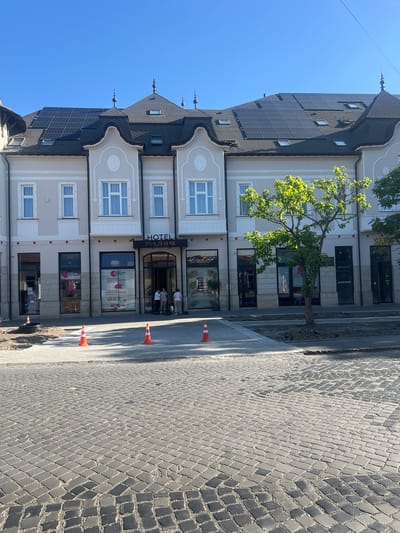 In Berehove, Ukraine, on a sunny day, a multi-story building with a light-colored facade and dark, mansard-style roofs is prominently featured. The building has several windows on its upper floors and a row of storefronts on the ground level. A sign above one of the storefronts reads "HOTEL PAVA DE LUXE". Solar panels are installed on the roof of the building. In front of the building, a tree with green leaves casts a shadow on the cobblestone street. Several orange traffic cones are placed on the sidewalk in front of the building, suggesting ongoing construction or maintenance. Three individuals are standing near the entrance of the hotel. The street is paved with cobblestones. The sky above is clear and blue.