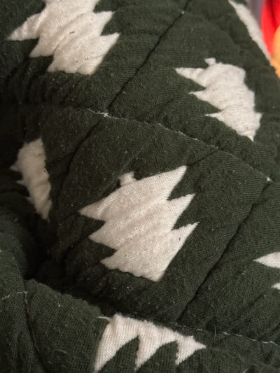 The image displays a close-up, overhead view of a quilted fabric with a repeating pattern of white, stylized Christmas trees on a dark green background. The fabric appears to be folded and creased, creating shadows and highlighting the texture of the quilting stitches. The pattern is consistent across the visible portion of the fabric. There are no people or any recognizable environmental features present in the image. The location in Santa Barbara, United States of America is not discernible from the provided image.