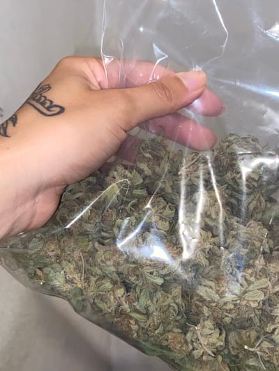 A person's hand holds a clear plastic bag filled with marijuana. The hand has a tattoo on the wrist. The bag appears full of dried flower buds. The scene's specific location within Monte Grande, Argentina, is indeterminable from the provided image.