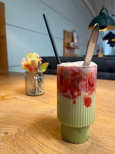 An indoor shot captures a matcha and strawberry drink in a fluted glass, positioned on a wooden table. The drink has a layered appearance, with green matcha at the bottom and a frothy, pinkish-red strawberry mixture on top. Two utensils, a black straw and a wooden spoon, are inserted into the drink. In the background, a small glass vase holds dried flowers. The wall behind the vase is painted a light blue with horizontal white trim. Two green pendant lights with gold interiors hang from the ceiling. A dark sofa is visible in the far background. The scene appears to be set in a cafe or restaurant. The image does not contain any specific landmarks or identifiers that would place it in Esslingen am Neckar, Germany, or any other specific city.