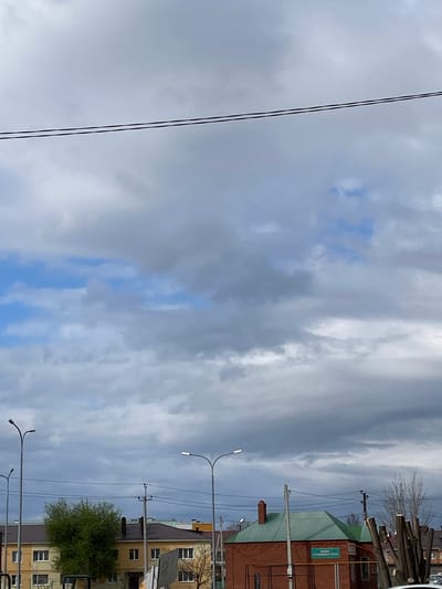 The image shows a street in Urussu, Russia. Overhead power lines stretch across the left side of the frame. On the right side of the frame are buildings, streetlights, and trees. The sky is overcast with a mix of gray and blue clouds.