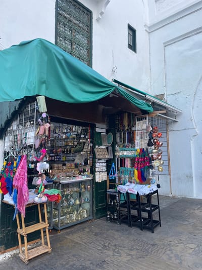 The image depicts a souvenir shop storefront located on a paved street in the city of Moulay Idriss Zerhoun, Morocco. The building exterior features white-washed stucco walls with visible wear and a decorative, dark green metal-grated window positioned above the storefront, along with a smaller, higher rectangular dark green-framed window to the right.
A dark green fabric awning extends from the building, covering the entrance area of the shop. The storefront is densely packed with merchandise. On the left, a metal grid displays numerous keychains, small purses, and accessories. Inside the shop, various shelves and racks hold small decorative items, including what appear to be bottles, metal crafts, and small mirrors. A glass display cabinet at the lower left contains multiple gold-colored, conical decorative objects and other metallic items.
To the right, strands of colorful beads and necklaces are hung vertically. Further right, colorful patterned textiles, possibly scarves, are displayed on a rack. Stacked silver-colored metal bowls or cups are visible on a display. On the sidewalk in front of the shop, a three-tiered light-toned bamboo-style wooden stand is positioned to the left. To the right, two black, three-tiered metal display stands are present; one holds folded white fabric items, while the other is empty. The ground is paved with grey, textured material. No individuals are present in the scene.