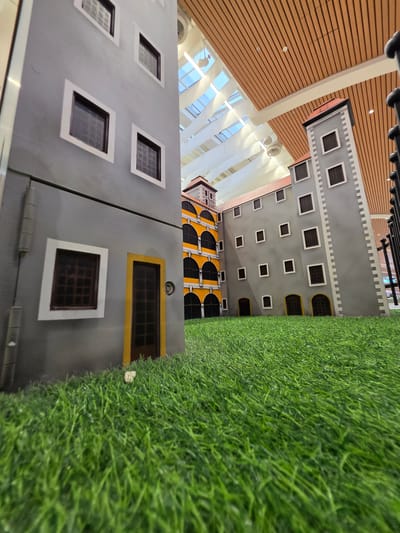 The image depicts a miniature architectural model of buildings with grey facades and dark-paned windows, set against a backdrop of a wooden-paneled ceiling and a skylight. The buildings are positioned on a green, artificial turf surface. In the foreground, the grass is in sharp focus, gradually blurring into the background. The scene appears to be indoors, possibly in a public space like a mall or exhibition center, given the skylight and ceiling structure. The style of the buildings suggests a historical or European architectural influence. There are no people or animals visible in the image. The location of Mladá Boleslav, Czechia is not determinable from the visual information provided in the image.