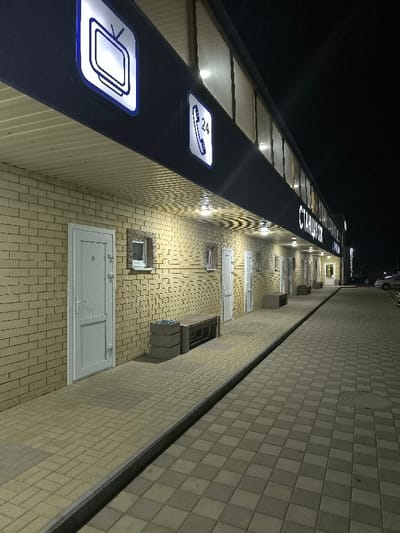 The image depicts the exterior of a commercial building at night in Raz"Yezd Vasil'yevskiy, Russia. The scene is illuminated by artificial light sources. The building features a light-colored brick facade and a prominent dark overhead structure extending outwards, forming a canopy. The underside of this canopy has a corrugated ceiling and is fitted with multiple recessed lights that cast circular patterns of illumination onto the brick wall and the paved ground below.

Above the canopy, three illuminated signs are visible. From left to right, the first displays a white retro-style television icon on a blue square background. The second shows a white telephone icon with the number "24" on a blue square background, indicating 24-hour service. Further to the right, a longer sign displays Cyrillic text, reading "СТАНЦИЯ-СТО" (Stantsiya-STO), illuminated in white against a dark background.

Along the brick wall, at least five identical white, solid doors with silver hardware are spaced evenly. Between some of these doors are small, rectangular windows with white frames, some appearing to have security grates.

A walkway paved with light-colored rectangular pavers runs parallel to the building. This walkway is separated by a dark grey curb from a wider paved area constructed of alternating light and dark grey square pavers. A circular utility cover is visible on this wider paved section in the foreground. Several dark-colored benches with concrete-like bases are positioned along the walkway in front of the building. Adjacent to each visible bench are square, stacked concrete or stone structures, possibly planters or waste receptacles.

No individuals or human activity are present in the image. In the distant right background, several parked light-colored vehicles are discernible, suggesting an adjacent parking area. The sky is dark, indicating nighttime conditions.