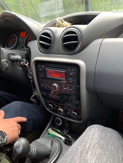 The image shows the interior of a car, likely a Dacia Duster, viewed from the driver's perspective. A person's hand, wearing a watch and a ring, is visible in the lower right corner, resting on their dark blue pants. The dashboard features the car's radio display, showing "FM1-P2 NOVOE," along with various buttons and knobs for controlling the audio system and climate. Two air vents are also visible on the dashboard. The gear shift is prominently displayed, indicating a manual transmission. The scene appears to be set during the daytime, with natural light filtering into the car, suggesting an outdoor setting, possibly in a rural area given the location context of Мядзельскі сельскі Савет, Belarus. The overall impression is one of a mundane car interior, with the focus on the functional elements of the vehicle and a hint of the driver's presence.