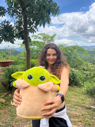 A young woman with long, curly brown hair stands outdoors, holding a large, lime-green plush toy resembling a baby Yoda. She is wearing a dark bikini top and black athletic shorts with a white wrap. Her arms are extended forward, cradling the toy. The background features lush green vegetation, rolling hills covered in trees, and a cloudy blue sky, suggestive of a mountainous or elevated rural landscape, possibly in Sơn Điền, Vietnam, known for its natural beauty. A tree trunk and foliage are visible to her left, and a large brown pot is also present on the left side of the frame.