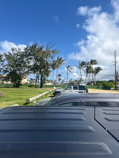 The image depicts a scene in Laie, United States, characterized by a clear blue sky with scattered white clouds above palm trees. A gray vehicle dominates the foreground. Adjacent to the gray vehicle and behind the fence, sits a white vehicle facing the same direction. A fence is visible. A grassy lawn and a road lined with trees and utility poles are present. A few buildings are noticeable in the background. The scene is outdoors and taken during daylight hours.