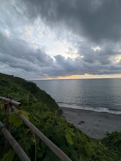 This is a wide shot taken from a grassy, vine-covered cliff overlooking a rocky beach and the sea in Tsikhisdziri, Georgia. The sky is filled with dramatic, dark gray clouds, with a sliver of yellow light visible near the horizon where the sun is setting or rising. The sea is a deep blue-gray, with small waves lapping at the shore. Two people are walking on the pebble beach, their figures small against the vastness of the landscape. In the foreground, a rustic bamboo fence is partially visible, covered in lush green vines.