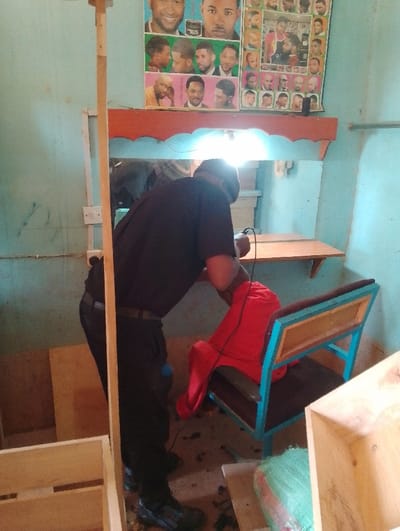 The scene is a barber shop in Ihururu, Kenya, located at latitude -0.389 and longitude 36.895. A barber, dressed in black pants and a black shirt, is cutting the hair of a seated customer wearing a red shirt. The customer is partially visible, sitting on a blue and black metal chair. The barber is using a handheld electric clipper. The shop's walls are painted light blue.  A mirror is mounted on the wall behind the barber and customer, with a bright overhead light illuminating the space.  In the background, wooden crates and a green sack are visible.  On the wall, behind the mirror, is a collection of framed photographs, likely displaying various hairstyles.