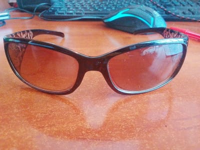 The image shows a pair of sunglasses with brown lenses and a black frame. The sunglasses are lying on a wooden table with an orange-brown textured surface. In the background, a computer keyboard and a computer mouse with a blue illuminated accent are visible. A red pen is also present to the right of the sunglasses. The lighting suggests it could be daytime, indoors. The scene appears to be a desk setup, possibly in a home or office environment. There is no discernible text in the image. The overall impression is of a casually placed personal item on a workspace.