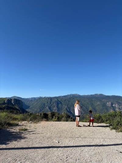 Two individuals, an adult and a child, stand on a gravelly overlook in Kućišta, Montenegro, facing a vast mountainous landscape. The adult, wearing a light pink t-shirt and dark shorts with a white bandage on their left leg, holds a mobile phone. The child, in a white patterned t-shirt and red shorts, stands to the right of the adult. They are positioned on a plateau covered in small stones and sparse vegetation, with a clear blue sky above. The background features a series of lush green mountains with rocky outcrops, indicating a natural, undeveloped area. The overall scene captures a moment of quiet observation of the expansive natural beauty of the region.