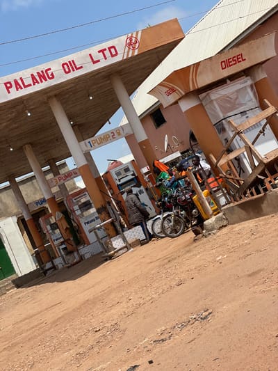 The image shows a gas station with the name "PALANG OIL LTD" prominently displayed. Several gas pumps are visible, with labels indicating "1 PUMP 2", "3 PUMP 4", and "PUMP". The word "DIESEL" is also visible on an awning. There are at least three motorcycles parked near the pumps, and several people are gathered around them. One person is wearing a dark jacket, another a green shirt, and a third a white shirt. A person in the background appears to be wearing a yellow jacket. A large yellow container, possibly for fuel, is placed on the ground next to a motorcycle. The ground is unpaved and appears to be dirt or gravel. The sky is blue with some clouds. The gas station appears to be in a somewhat rural or less developed area, likely within the city of Tangu, Nigeria, given the context.