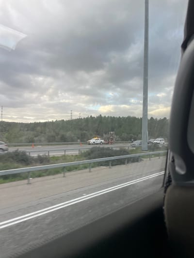An overcast sky looms over a multi-lane highway, viewed from inside a vehicle. A line of trees borders the road. A white service vehicle with flashing lights is parked on the shoulder, attending to a vehicle being towed. Orange cones delineate a work zone on the shoulder of the highway. Transmission towers are visible in the background along the tree line.