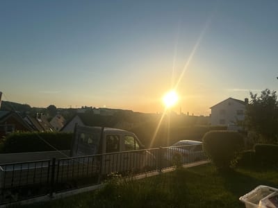 The image depicts a residential area in Mainburg, Germany, during a sunset. The sun is low in the sky, casting a warm, golden light and creating lens flare. In the foreground, there is a grassy lawn with a black metal fence. Beyond the fence, a flatbed truck is parked. Further back, several houses with pitched roofs are visible, along with a white building on the right. The background shows a more distant cityscape with buildings and trees, all bathed in the soft light of the setting sun. The sky transitions from a bright yellow near the horizon to a clear blue at the top of the frame.