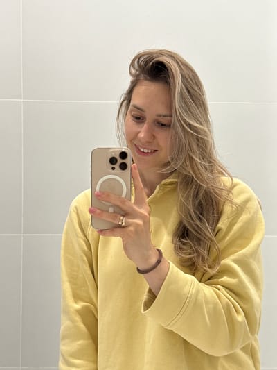 This mirror selfie, currently rotated 90 degrees counter-clockwise, features a smiling woman with long, wavy light brown hair. She is wearing a mustard yellow long-sleeved top, possibly a sweatshirt. On her left wrist, she has a dark brown beaded or woven bracelet, and on her right ring finger, she wears two rings. Her fingernails are painted a soft pink.
The woman is holding a light gold or champagne-colored smartphone, likely an iPhone, encased in a clear protective cover. A distinctive white circular MagSafe ring is prominent on the back of the phone, partially obscuring the Apple logo. Her gaze is directed towards the phone screen, indicating she is actively taking the picture.
The background consists of light-colored, possibly white, tiled walls with visible grout lines, suggesting an indoor setting such as a bathroom or a modern room. The lighting appears bright and even. The overall impression is one of casualness and a pleasant mood, conveyed by her gentle smile. The provided location context for this media is Новая Казмаска, Russia. No specific time of day or weather is discernible.