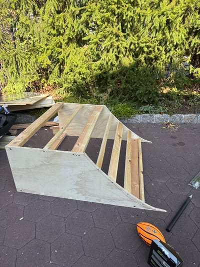 The image depicts a partially constructed wooden skate ramp or jump situated outdoors on a patio with dark hexagonal pavers in Cresskill, United States. The ramp is composed of light-colored plywood panels and lighter-toned lumber framing, secured with visible fasteners. Pencil markings are present on the plywood surfaces, indicating construction lines.

To the left of the ramp, against a dense green evergreen hedge, a pile of additional building materials is visible, including multiple sheets of plywood, lumber pieces, and a folded dark fabric object. The hedge forms the background, consisting of numerous coniferous trees with lush foliage.

To the right of the ramp, on the paver surface, several items are scattered: an orange Nerf-brand football, a partially visible cardboard box labeled "DeckFast" (likely containing construction fasteners), a black utility knife with a blade guard, and a black cylindrical object, possibly a tool handle or component. No individuals are present, and no active construction or human interaction is depicted.