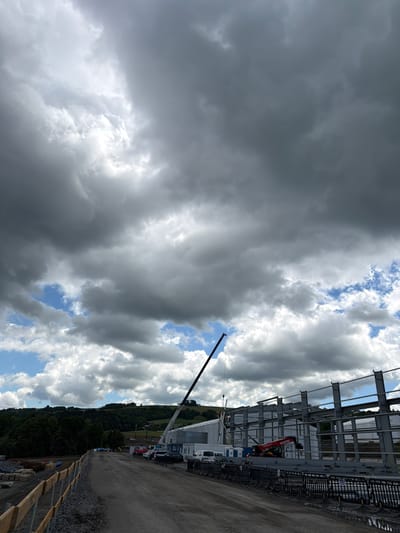 The image shows a construction site in Jodłownik, Poland, under a dramatic, cloudy sky. A large metal structure, presumably a building in progress, is visible on the right, with steel beams forming its frame. A crane with a long boom extends into the sky from the left, and a red Manitou telehandler is positioned near the building. Several vehicles, including cars, vans, and portaloos, are parked on a dirt road leading into the site. In the foreground, a gravel path is bordered by a wooden fence. Behind the construction, rolling green hills covered in trees form the backdrop, suggesting a rural or semi-rural setting. The sky is filled with heavy, dark clouds interspersed with patches of blue sky and brighter clouds, indicating potentially unstable weather. There are several construction workers visible on the scaffolding of the building.