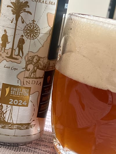 A close-up shot shows a beer can and a glass of amber-colored beer with a thick head of foam. The beer can, which is partially visible, has a design that includes a map, an elephant, and two figures. Text on the can indicates "INDIA", "FINEST BEER SELECTION", and "2024". The background is out of focus and appears to be a textured surface. The image is taken in Mohyliv-Podilskyi, Ukraine.