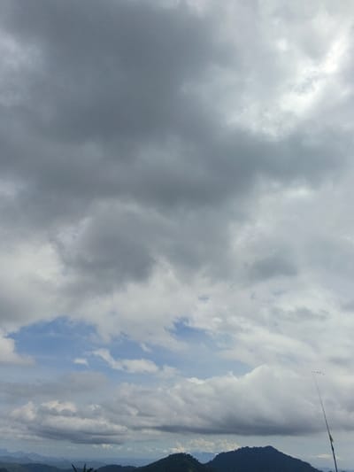 The image depicts a mountainous landscape under a partly cloudy sky in Ngọc Tụ, Vietnam. The majority of the frame is filled with an expansive sky, comprising various shades of grey and white stratocumulus clouds, with intermittent patches of clear blue sky visible, primarily in the lower-middle section. In the lower third of the image, multiple layers of dark green, forested mountains extend across the horizon, appearing hazy in the distance. A slender, dark pole or stick with a small yellow band near its visible base extends diagonally upwards from the bottom-right corner, terminating with a small, indistinct object at its tip and a faint dotted line stretching horizontally to the right. Minimal dark green foliage is visible along the extreme bottom edge of the frame. No individuals, animals, or explicit actions are discernible.