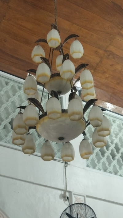 A multi-tiered chandelier with many individual light fixtures hangs from a wooden ceiling. Each light fixture has a white, frosted glass shade with horizontal yellow bands. The chandelier has a central, tiered body and several arms extending outwards, each supporting multiple lights. The background shows a white wall with a decorative, translucent screen or window, and a portion of a black oscillating fan is visible at the bottom of the image. The scene appears to be indoors.