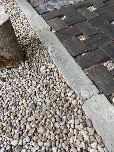 The image shows a close-up, high-angle view of a textured outdoor surface in the vicinity of Лышчыцкі сельскі Савет, Belarus. The ground is composed of a variety of materials. To the left, a rough, weathered tree trunk segment is visible, with bark and some moss. Adjacent to the trunk is an area covered in small, multi-colored pebbles and gravel. A concrete border, rough and somewhat broken, runs diagonally across the middle of the frame, separating the pebbles from a section of dark wooden planks laid in a herringbone pattern. These wooden planks appear to be weathered and are interspersed with small stones and gravel in the gaps between them. The overall impression is of an aged, rustic pathway or garden feature. There are no people or animals present in the image.