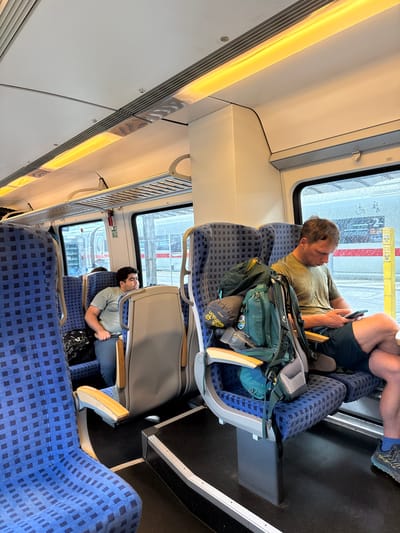 The image depicts the interior of a train carriage with two men seated. The man on the right is wearing a green t-shirt and dark shorts, and is looking down at his smartphone. He has a large green backpack with various items attached, resting on the seat next to him. The man on the left is wearing a grey t-shirt and dark pants, and appears to be resting his head against the window. The train seats are upholstered in blue fabric with a dark blue checkered pattern. The carriage is illuminated by warm-toned lights along the ceiling. Outside the window, a train platform is visible, with some rain or condensation on the glass. The scene appears to be in Bitterfeld-Wolfen, Germany, as indicated by the context provided.