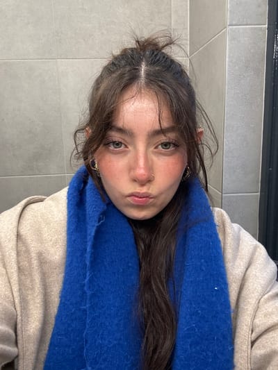A young woman with fair skin, rosy cheeks, and green eyes gazes at the viewer. Her dark hair is partially pulled back, and she has a nose ring and delicate earrings. She is wearing a beige jacket and a bright blue, fuzzy scarf. The background is a neutral gray, suggesting a tiled wall. The photo is slightly rotated.