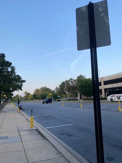 A wide-angle outdoor shot captures an empty parking lot adjacent to a sidewalk in Derwood, Maryland. The sky is clear and blue, with a few wispy clouds and faint red specks visible in the distance, possibly from fireworks or lights. A tall, black signpost with a blank white sign faces the viewer, positioned on the right side of the frame. The parking lot is lined with yellow bollards and marked parking spaces. A dark-colored SUV is parked in the lot, and a Coach USA bus is visible further back, parked in front of a multi-level parking garage. On the sidewalk, two young individuals are seated, appearing to be resting or engaged with their phones. Trees line the perimeter of the parking lot and sidewalk, with a streetlight visible on the left. The scene appears to be set in the late afternoon or early evening, given the soft lighting.