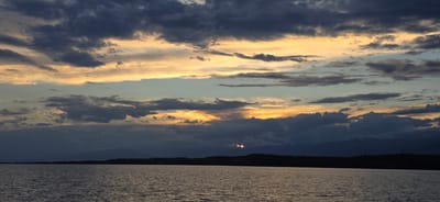 A wide-angle shot captures a serene sunset over a calm body of water, likely Issyk-Kul Lake near Pristan'-Przheval'sk, Kyrgyzstan. The sky is a dramatic canvas of dark, heavy clouds interspersed with streaks of warm yellow and orange light where the sun is setting. These colors illuminate the undersides of the clouds, creating a textured and moody atmosphere. Below the vibrant sky, a dark, silhouetted treeline stretches horizontally across the horizon, with faint mountain ranges visible in the distance. The water in the foreground is a deep, muted gray, reflecting the subdued light of the sky with gentle ripples. No people or specific man-made structures are clearly visible, contributing to the natural, tranquil feel of the scene.