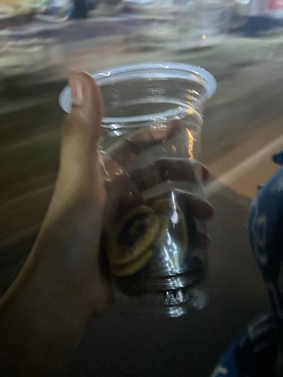 An out-of-focus, close-up shot captures a hand holding a clear plastic cup filled with small, round objects, possibly cookies or candies, in Walennae, Indonesia. The cup is partially obscured by the hand. The background is blurred, with hints of streetlights and foliage, suggesting an outdoor setting at dusk or night. The overall lighting is dim, with a slight yellow-orange hue.