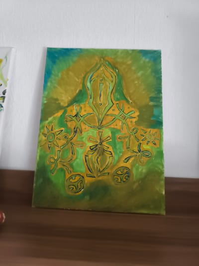 An abstract painting with yellow and green hues is displayed on a wooden shelf against a white, textured wall. The painting features a central, symmetrical design with flowing lines and circular motifs, outlined in dark blue. To the left of the painting, a portion of another artwork, also featuring white and green with yellow accents, is visible. The scene is illuminated by natural light, casting soft shadows. The location is Eitorf, Germany.