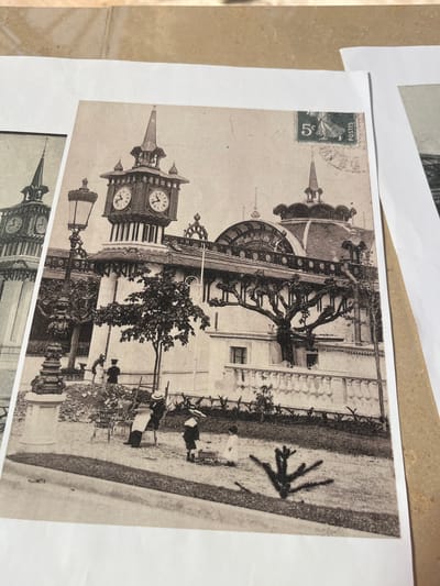 The sepia-toned photograph depicts a street scene in Évian-les-Bains, France, featuring a grand building with a prominent clock tower. The building has multiple windows and a decorative roofline with ornate railings. A large, stylized tree with bare branches is positioned in front of the building. In the foreground, several people are present. A man in a hat and a child in a sailor suit are near a seated figure. There are also chairs scattered on the ground. A streetlamp stands to the left of the clock tower. A postage stamp with the denomination "5" and the word "POSTES" is visible in the upper right corner of the photograph, indicating it is likely a postcard. The overall scene suggests a public space or a notable landmark in the city.
