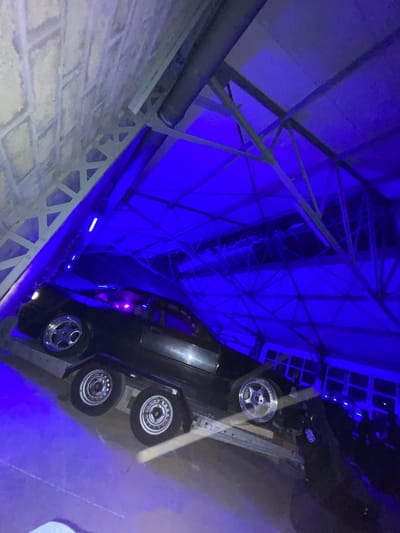 This media file captures an artistic installation within a large indoor space, likely a gallery or cultural venue in Vitry-sur-Seine, France. The central subject is a dark-colored car, possibly an older model, mounted on its side or at an acute angle against a metal framework. The car's front wheels feature distinctive shiny, multi-spoke chrome rims, while the visible rear wheels appear to have steel rims with hubcaps. Inside the car, a subtle purple or pink glow emanates from interior lights.
The entire scene is dramatically illuminated by intense artificial blue lighting, which casts a cool, mysterious ambiance. This blue light highlights the intricate network of metal trusses and beams forming the ceiling structure above the car. Linear blue light strips also line the metal support structure beneath the car, drawing attention to its suspended position. To the left of the installation, a textured brick wall is visible, providing a contrasting element to the industrial structure and vibrant lighting. The perspective is an upward-looking shot, emphasizing the height and unconventional placement of the car. No discernible text is visible on the car or surrounding structures. The artificial lighting suggests an indoor setting, likely during the evening, designed to enhance the artistic presentation.