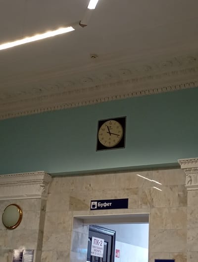 The image depicts an interior scene, likely a public building such as a train station or airport, given the presence of a clock and signage. A circular clock with Roman numerals is prominently displayed on a light blue wall. Below the clock, a dark blue sign with white text reads "Буфет", which translates to "Buffet". To the left of the buffet sign, there is a circular mirror with a gold frame mounted on a marble column. Further down, partially visible, is an entrance to a room with a sign reading "ПК-2" and some additional text in Cyrillic that appears to be emergency contact information. The ceiling features decorative molding and fluorescent lighting. The floor appears to be made of marble or a similar polished stone. There are no people visible in the frame. The image does not contain any specific visual cues to definitively place it in Kalinkavichy, Belarus; this information is based on external knowledge.
