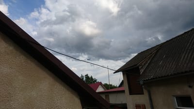 This image depicts a residential scene in Zhnyborody, Ukraine, under a cloudy sky. The foreground is dominated by the angled roofs and textured walls of houses. The roof on the left is a light beige stucco, with a dark red, sloped metal trim. The roof on the right is covered in corrugated, weathered grey material, suggesting an older structure. A dark power line runs diagonally across the frame, passing in front of the buildings.
In the middle ground, several other buildings are visible, including one with a bright red roof and another with white siding. Trees are scattered behind these structures, indicating a semi-rural or suburban environment.
The most prominent detail in the sky is a small airplane flying in the distance, silhouetted against the dramatic, overcast clouds. The clouds are a mix of dark grey and lighter white, creating a dynamic and somewhat dramatic atmosphere. The presence of the airplane suggests it is daytime, likely late afternoon or early evening given the cloud cover. The weather appears to be overcast, possibly with a threat of rain. There is no visible text in the image. The overall impression is one of everyday life in a Ukrainian village, with the passing of an airplane adding a touch of the modern world to the rural landscape.