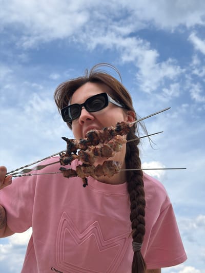 The image depicts a woman holding three skewers of grilled meat against a backdrop of a partly cloudy blue sky. The woman wears large, dark sunglasses and a light pink sweatshirt with a geometric design. Her hair is styled in a braid. The skewers are positioned close to the camera, slightly obscuring her face. The meat appears charred in some areas. There is no information to identify the location as Zīvartiņš, Latvia.