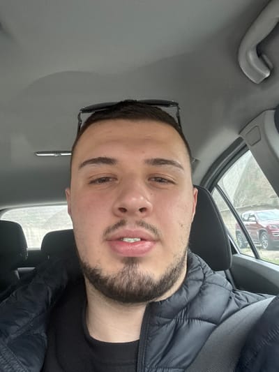 A man is seated in a car. He is wearing a black quilted jacket and a black t-shirt. Sunglasses are perched on the side of his head. He has dark hair and a beard. The interior of the car is gray. A car can be seen outside the window. The location is Byala Reka, Bulgaria.
