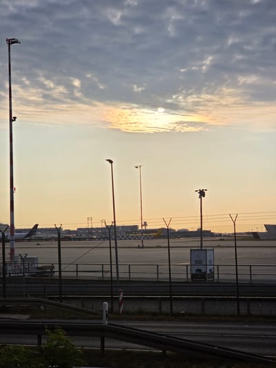 An exterior, eye-level, vertical, medium shot captures the Frankfurt Airport in Kelsterbach, Germany, at dusk. The sky is a gradient of muted blues and pale oranges, with scattered, textured clouds. A partially obscured sun shines through the clouds, casting a soft light. In the foreground, a chain-link fence with barbed wire topping separates the viewer from an airport tarmac. Several tall, red and white striped poles, likely for lighting or marking, stand prominently. One pole on the far left has a red light at its top. In the middle ground, several airplanes are parked, with one visible tail featuring the Lufthansa logo. In the background, airport buildings and structures are visible. Two small signs on the fence in the middle ground read "WATCH TOWER." A road with guardrails is in the immediate foreground, with some green foliage to the left. The overall scene depicts a tranquil, yet active, airport environment at the end of the day.