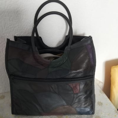 A dark, patchwork leather tote bag with two rounded handles is shown against a white, textured background. The bag features a variety of dark leather pieces stitched together in a mosaic pattern, with some sections appearing as rounded or curved shapes. A horizontal zipper divides the lower portion of the bag, which also displays the patchwork design. The bag is upright and appears to be empty. To the right of the bag, a portion of a yellow object, possibly a cushion or a rolled-up textile, is visible. The lighting is neutral, suggesting an indoor setting. There is no visible text in the image.