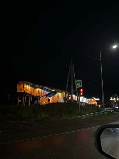 This image depicts a pedestrian overpass illuminated by warm orange and cool blue lights at night. The overpass is situated alongside a road, with a guardrail visible in the foreground. A traffic light with a red signal and a pedestrian crossing sign are present near the overpass. The sky is dark, indicating nighttime. The scene appears to be an urban or suburban environment, possibly near a road intersection or transportation hub.