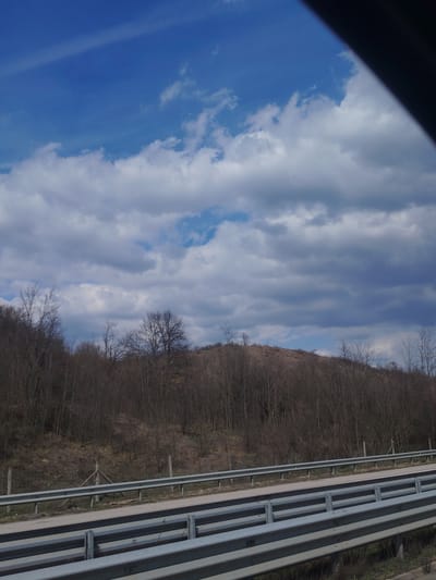 The scene depicts a rural highway landscape located in Ravnishte, Bulgaria, observed from an elevated position within a vehicle, indicated by a dark diagonal obstruction in the upper right. The sky overhead is partially cloudy, featuring patches of clear blue in the upper left, crossed by a faint white contrail. Below this, an expanse of white cumulus clouds transitions to darker grey stratocumulus clouds stretching across the horizon. The mid-ground is dominated by a dense treeline of bare deciduous trees, indicating a season without foliage, covering a gentle hillside. Beyond the treeline, the upper slope of the hill is visible, covered in sparse, dry, light brown vegetation. In the immediate foreground, a multi-tiered configuration of galvanized steel highway crash barriers (guardrails) extends diagonally across the lower portion of the frame. Behind the crash barriers, a strip of dry grass and soil is visible, followed by a wire fence with wooden posts, one of which is reinforced with a triangular wooden support structure. No individuals, discernible actions, or interactions are present in the image.