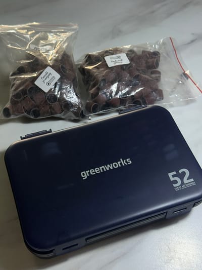 The image displays a dark blue accessory case and two clear plastic bags filled with sanding bands, all placed on a light-colored, possibly marble surface.
The rectangular, dark blue case, likely made of plastic, features the brand name "greenworks" printed vertically in white on its side. At the top right, the number "52" is prominently displayed in white, accompanied by smaller text that reads "LUCKY ACCESSORIES" and "SMART AV ELECTRIC ROTARY TOOLS," indicating its purpose for storing rotary tool accessories.
Next to the case are two transparent, resealable plastic bags, each containing a quantity of small, brown, cylindrical sanding bands or sleeves. These are commonly used with rotary tools for sanding or grinding. Both bags have white rectangular stickers. The sticker on the upper bag partially shows "Спасибо за покупку" (Thank you for your purchase) and a five-star rating graphic with "faniform of". The sticker on the lower bag clearly displays "400" which typically denotes a grit size for abrasives, and also contains the partially visible "Спасибо за покупку" and five-star rating.
The overall scene suggests an indoor setting with bright, even lighting. There are no people present, and the image focuses solely on these items. The objects appear to be new or well-maintained, implying readiness for use or storage.