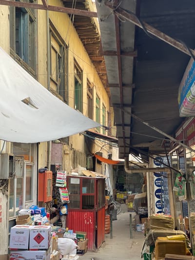 The image depicts a narrow alleyway in a bustling market in Kabul, Afghanistan. The buildings lining the alley are old, with weathered yellow facades and wooden window frames. A white canvas awning is stretched across the left side of the alley, providing shade. Various stalls and shops are set up along both sides of the alley, displaying a variety of goods.

On the left, there is a shop with glass windows, and a collection of boxes, including some with UN markings and hazard symbols, are stacked in front. A red metal cabinet and a small red kiosk are also visible.

On the right side of the alley, more shops are visible. Some have shelves stocked with various items, and there are signs for "NIVEA" products. Bags of goods are stacked high, and some items are hanging.

The alley floor is paved with concrete, and there are pipes and electrical wires visible overhead, indicating a functional but perhaps unkempt infrastructure. In the background, a bicycle can be seen, suggesting continued use of traditional transportation methods. The overall impression is one of a vibrant, working marketplace, characteristic of many urban areas in Kabul.