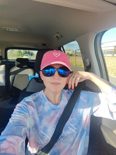 A woman with bright orange fingernails and a pink baseball cap with a white heart emblem is taking a selfie in the driver's seat of a car. She is wearing a tie-dye shirt with blue, pink, and white patterns. The woman is wearing sunglasses that reflect the surrounding environment, showing a blue sky and parts of the car interior. The car appears to be on a road, with trees and houses visible through the back window. The interior of the car shows dark gray upholstery on the seats and a black seatbelt. The scene takes place during daylight, indicated by the bright sunlight and shadows. The location is identified as Мядзельскі сельскі Савет, Belarus.