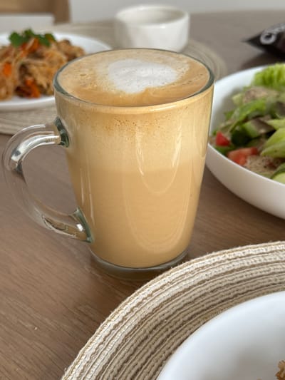 A close-up, eye-level shot captures a clear glass mug filled with a frothy, caramel-colored beverage, likely coffee or a latte, placed on a wooden table. The mug's handle is visible on the left. In the blurred background, to the left, is a white plate with food, possibly noodles and vegetables. To the right, a white bowl contains a salad with green lettuce, cucumber, and tomato. The scene is set in a dining environment, though no specific location in Sakmary, Russia, can be determined from the visual information.